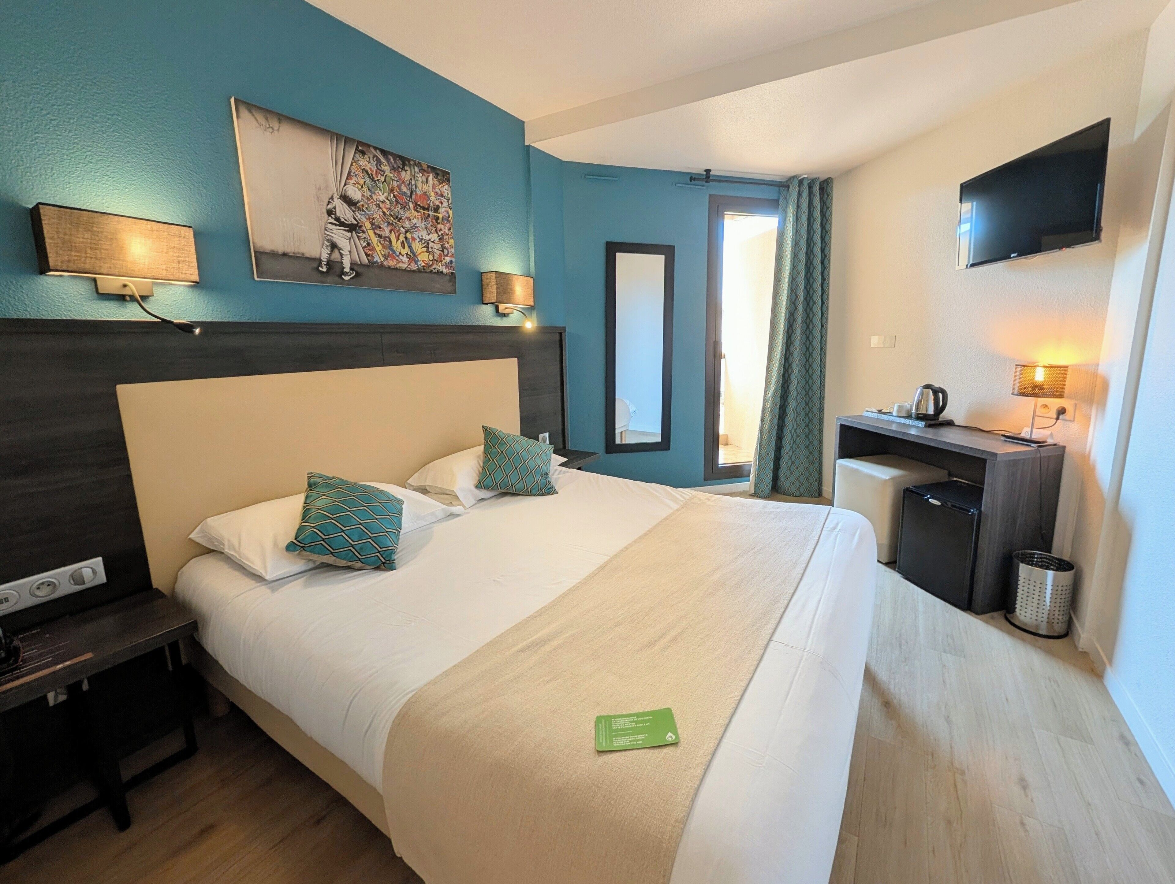 A hotel room with a bed, bedside table, lamp, artwork, desk, and TV.