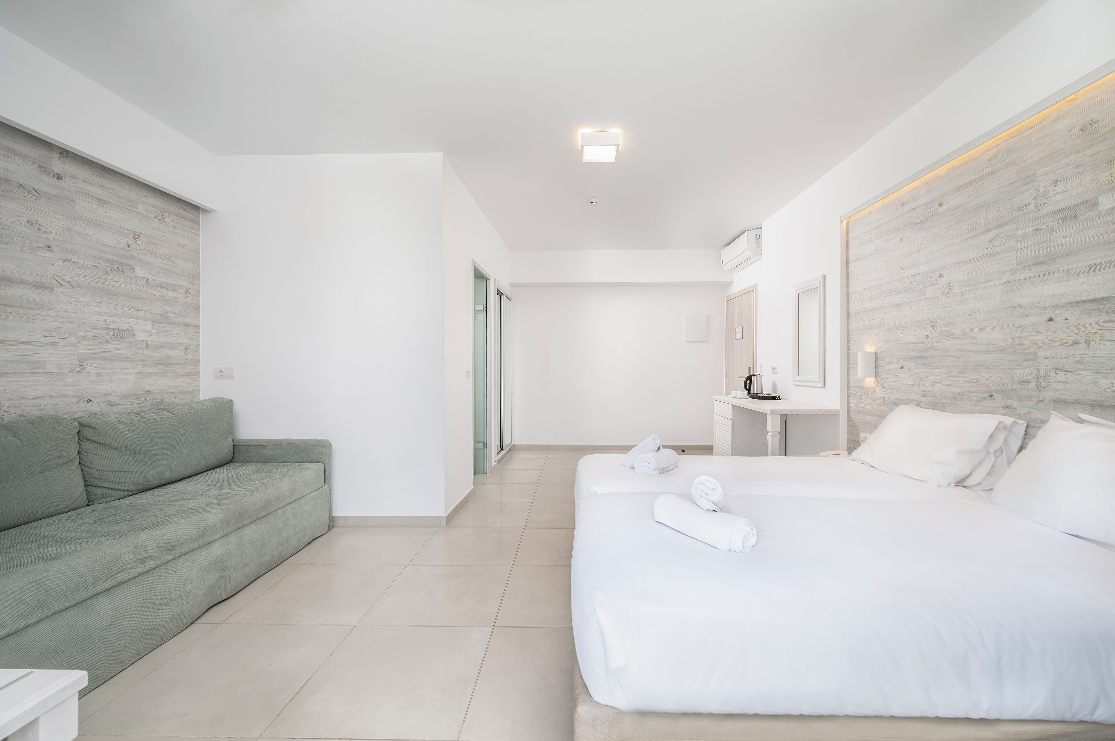 A modern hotel room with a grey sofa, a large bed with white linens, and a wooden accent wall.