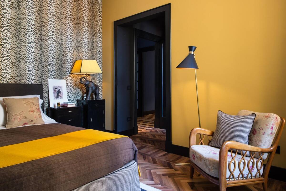 A hotel room with a bed, a chair, a lamp, a nightstand, and a patterned wallpaper.