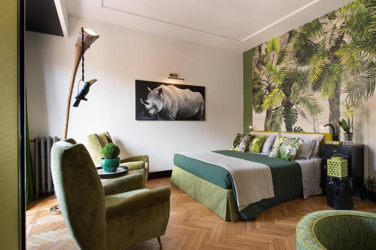 A bedroom with a bed, a chair, a wall-mounted lamp, a painting of a rhinoceros, and a tropical-themed mural.