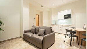 A modern living room with a sofa, a dining table with chairs, and a kitchen area with cabinets and a microwave. - Suites You Opera (Madrid)
