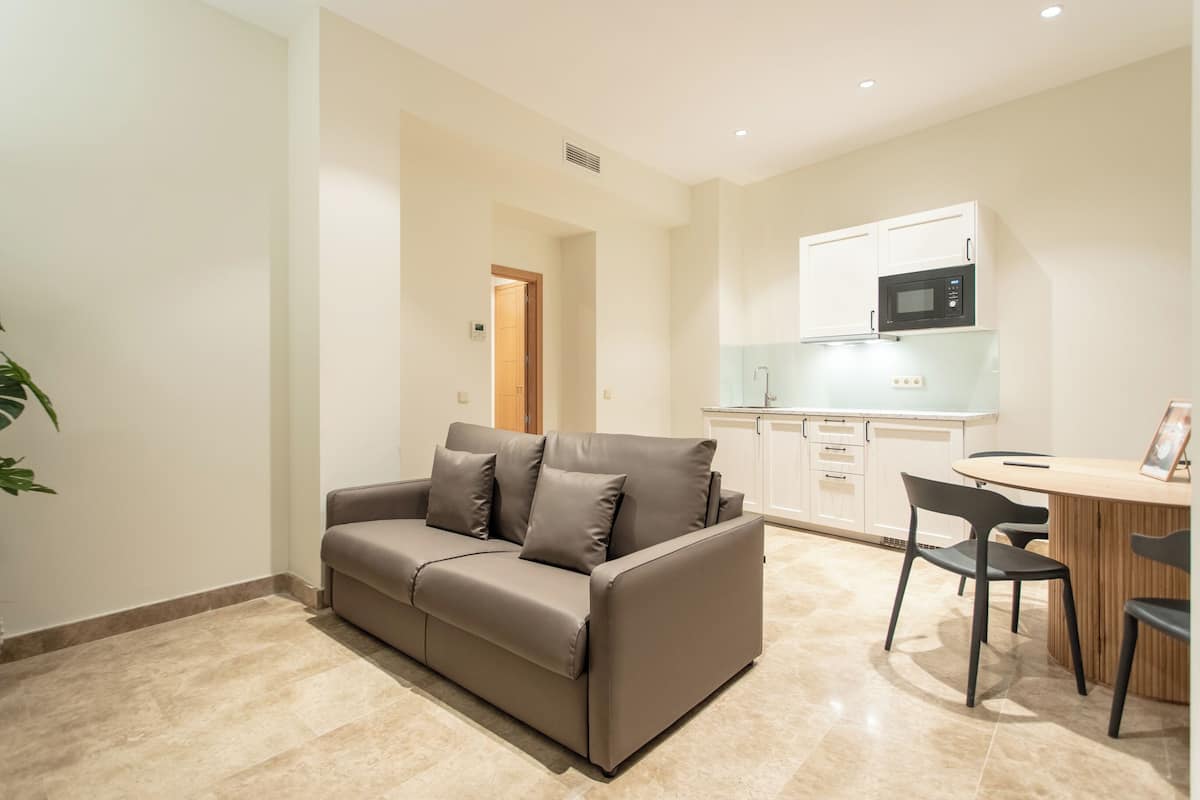 A modern living room with a sofa, a dining table with chairs, and a kitchen area with cabinets and a microwave.