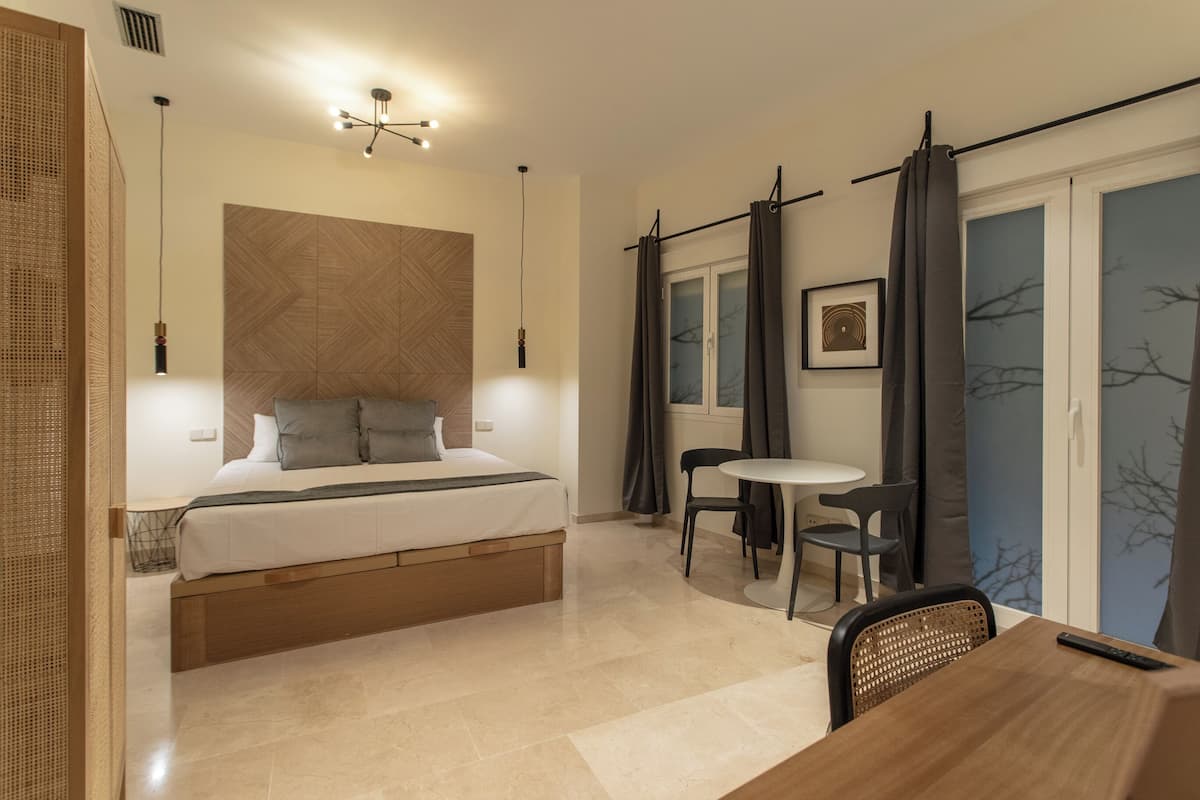 A modern hotel room with a bed, a small dining table, and a chair. There is a window with curtains, a picture on the wall, and a ceiling light fixture.