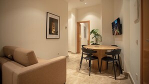 A modern living room with a beige sofa, a round wooden table with four black chairs, a framed picture on the wall, and a potted plant. - Suites You Opera (Madrid)