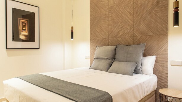 A modern bedroom with a wooden headboard, a large bed with a grey throw blanket, a framed picture on the wall, and a small round table with a wireframe base. - Suites You Opera (Madrid)
