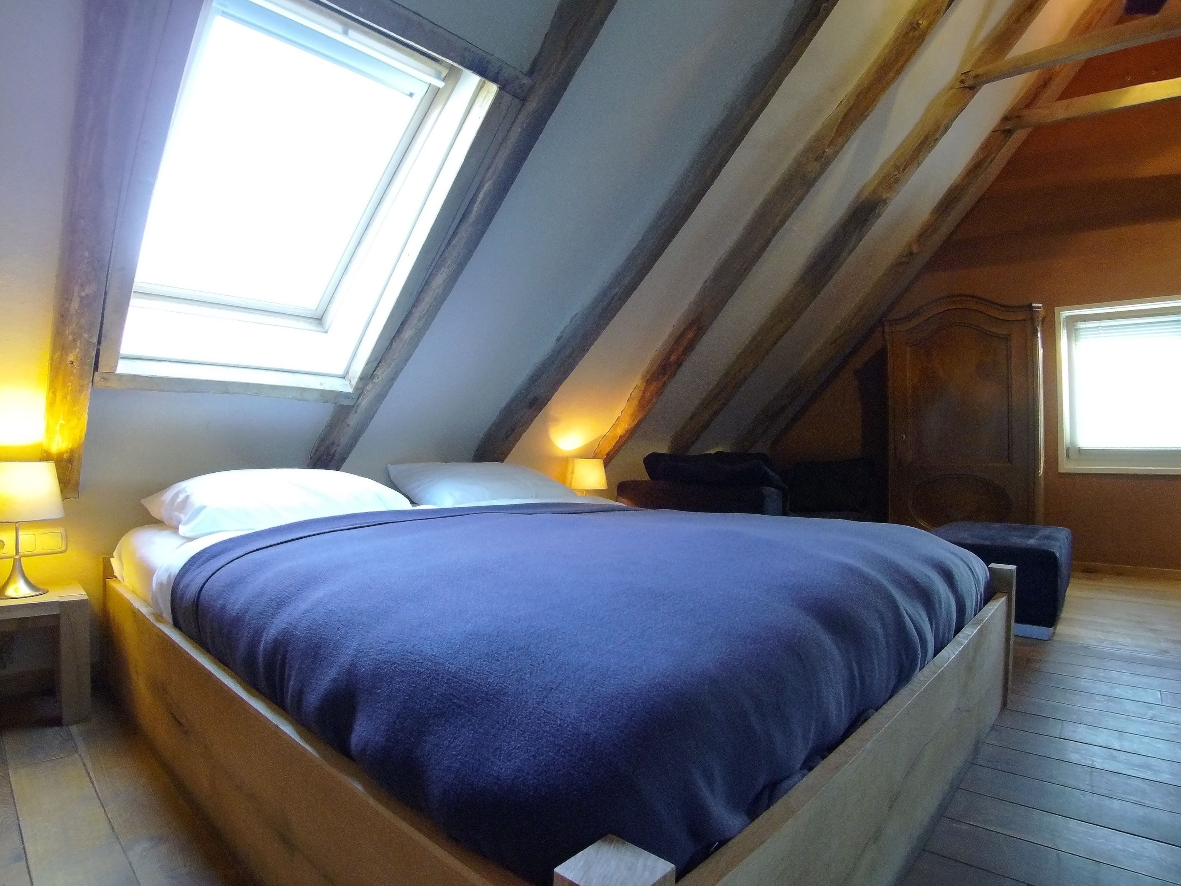 A bedroom with a sloped ceiling, wooden beams, a bed with a blue bedspread, a nightstand with a lamp, and a window providing natural light.