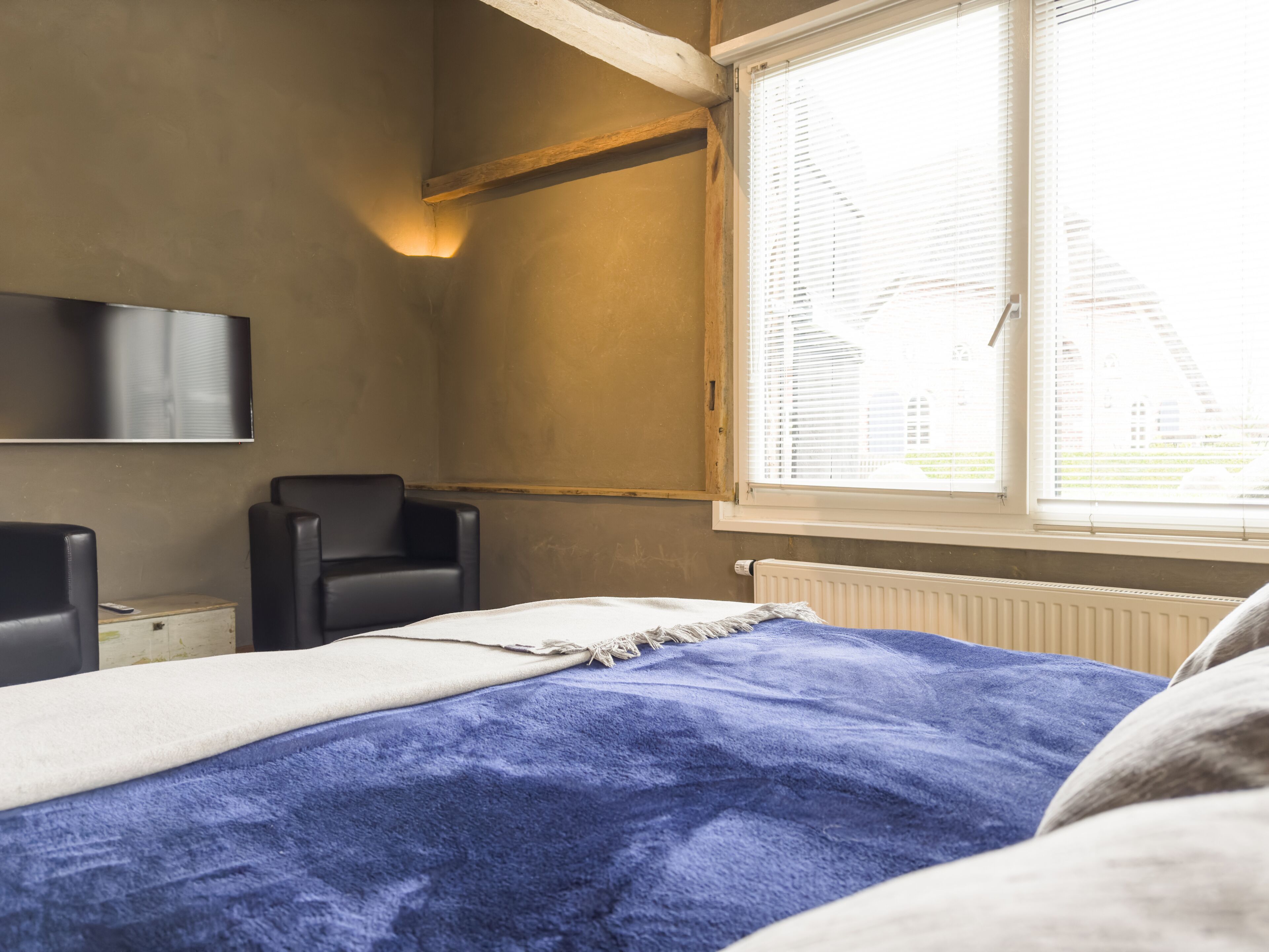 Une chambre à coucher comprenant un lit, une chaise, un téléviseur fixé au mur et une fenêtre avec des stores.