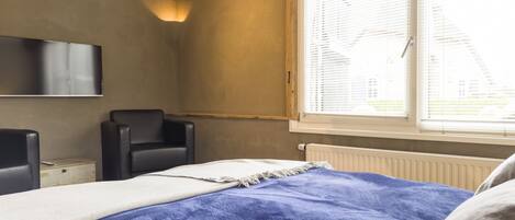 Une chambre à coucher comprenant un lit, une chaise, un téléviseur fixé au mur et une fenêtre avec des stores.