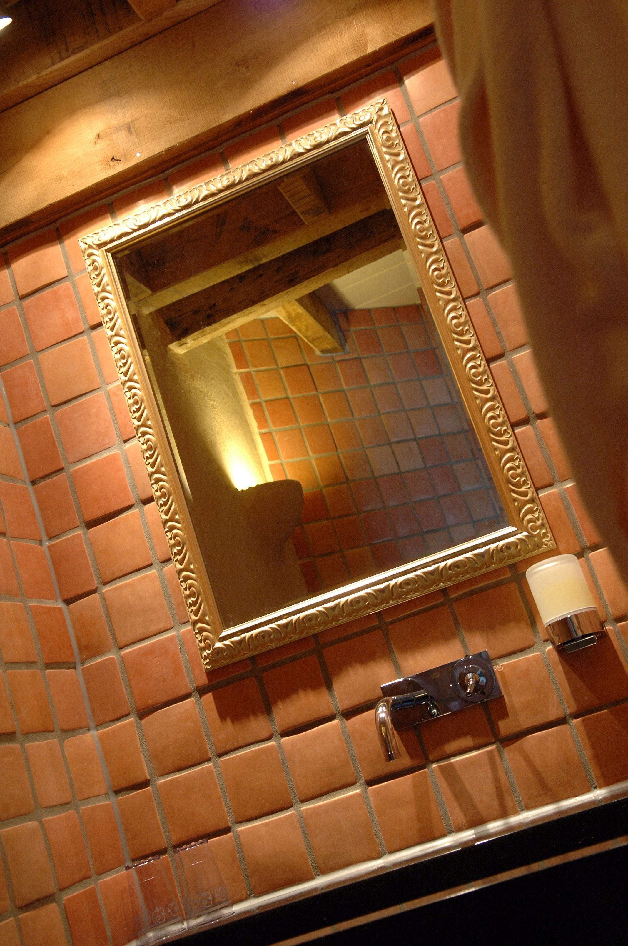 Une salle de bain avec un mur recouvert de carreaux, un miroir doté d’un cadre décoratif et un luminaire fixé au mur.