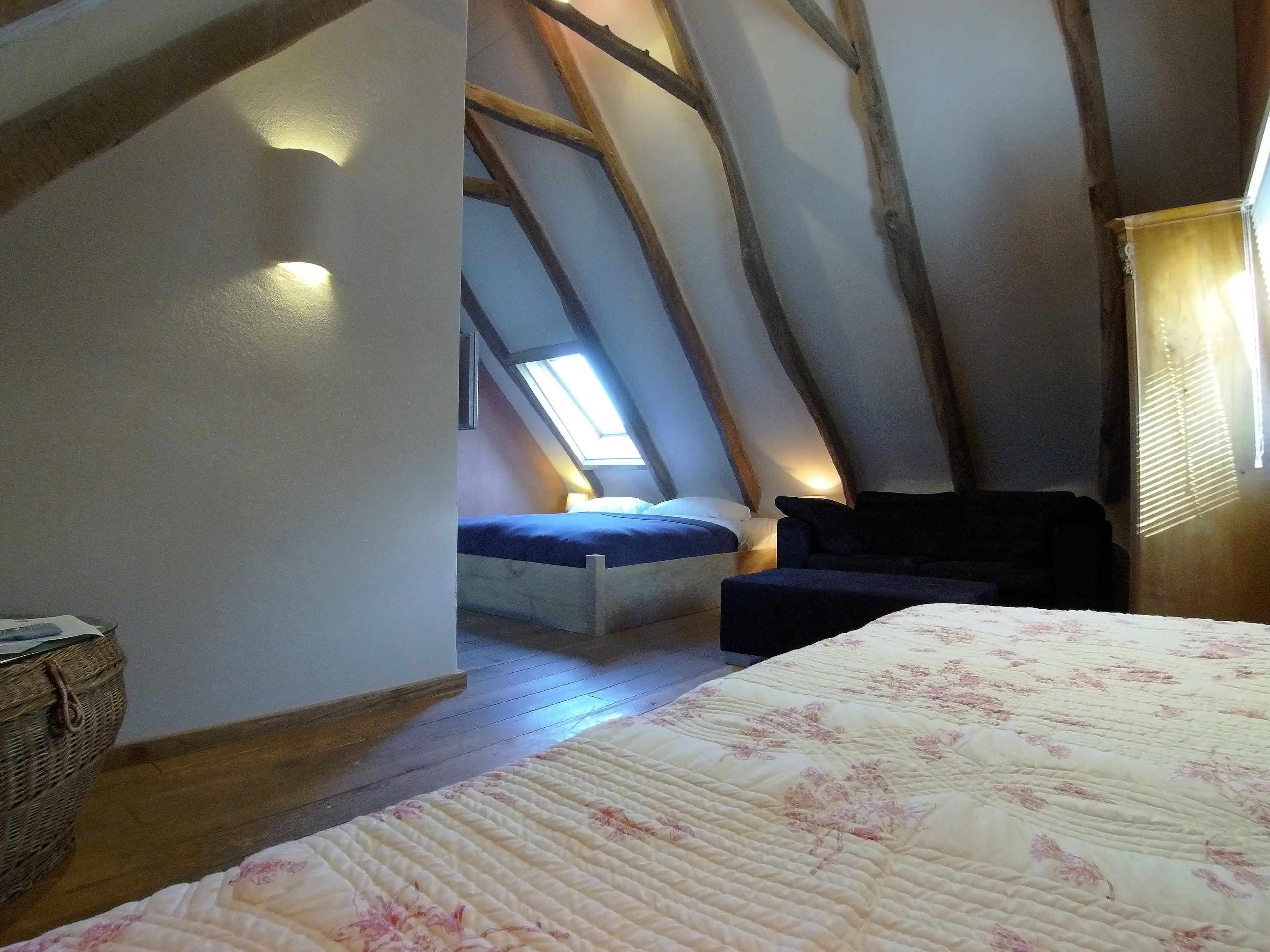 A bedroom with a sloped ceiling, a bed with a floral duvet, a small sofa, and a window providing natural light.