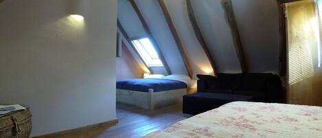 Une chambre avec un plafond en pente, un lit recouvert d’une couette à motifs floraux, un petit canapé et une fenêtre laissant entrer la lumière naturelle.