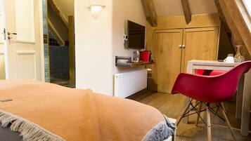 A bedroom with a bed, a red chair, a TV, a wooden wardrobe, and a kitchen area with a kettle and a coffee maker.