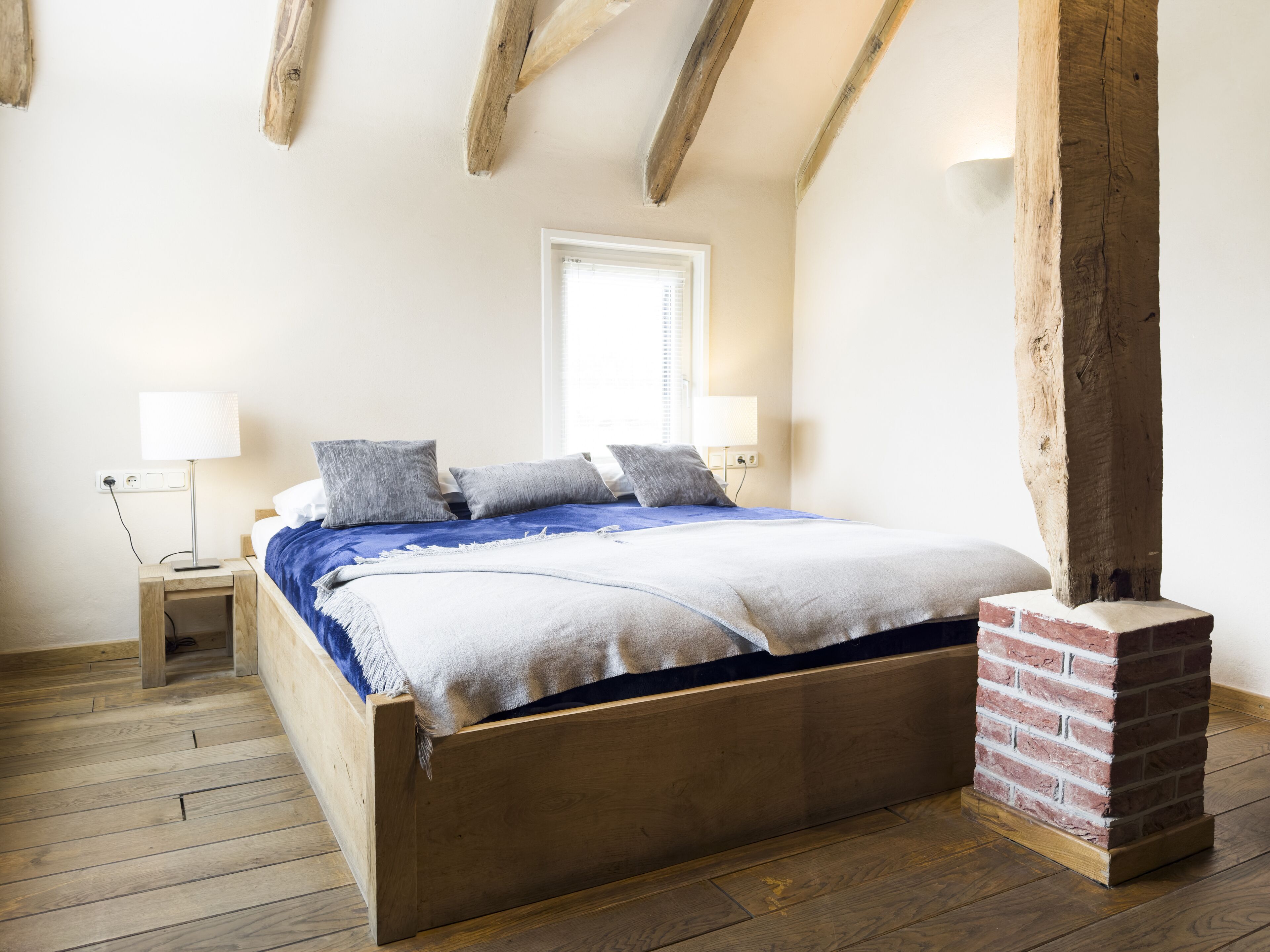 Quarto com cama de madeira, mesa de cabeceira, coluna de tijolos e janela com persianas.