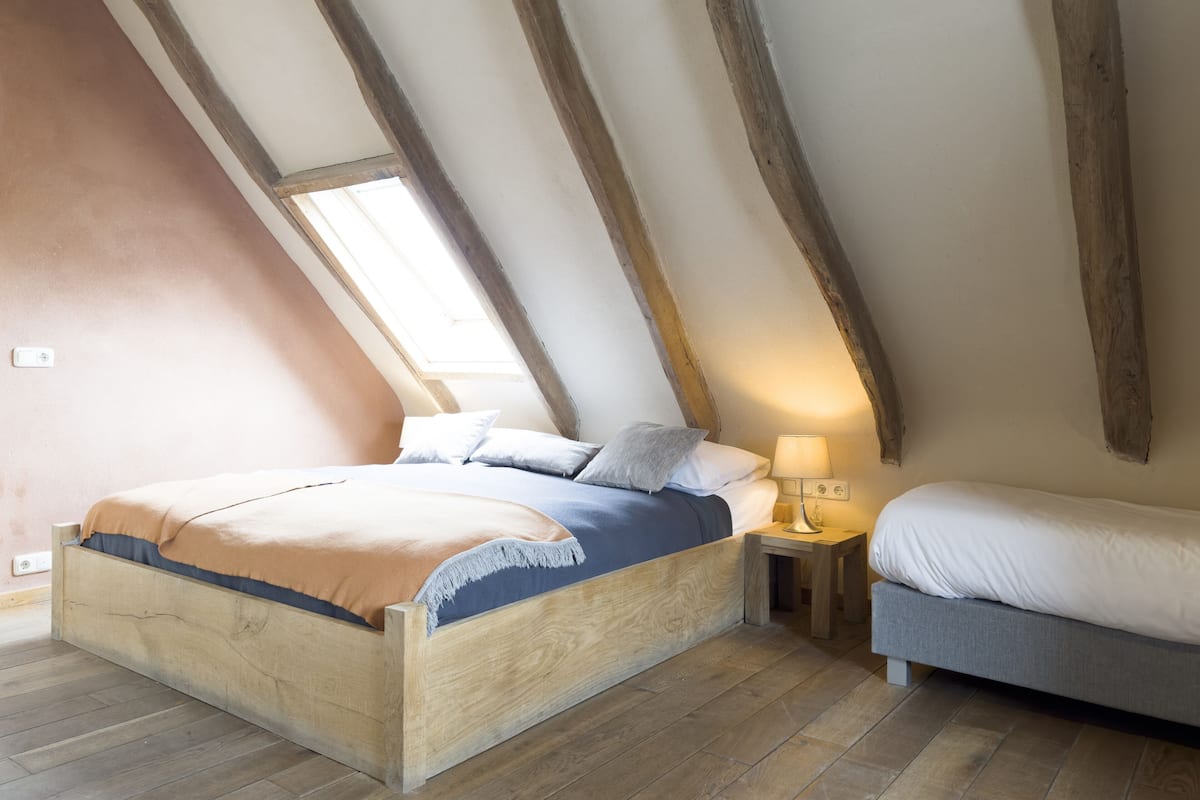 Une chambre avec un plafond en pente, des poutres en bois, un lit recouvert d’une couvre-lit bleue et orange, une table de chevet avec une lampe, et une petite table de chevet avec une autre lampe.