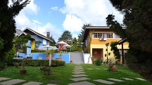 Un patio con un sendero de piedra que conduce a un edificio amarillo con puerta roja, rodeado de follaje y plantas en macetas.