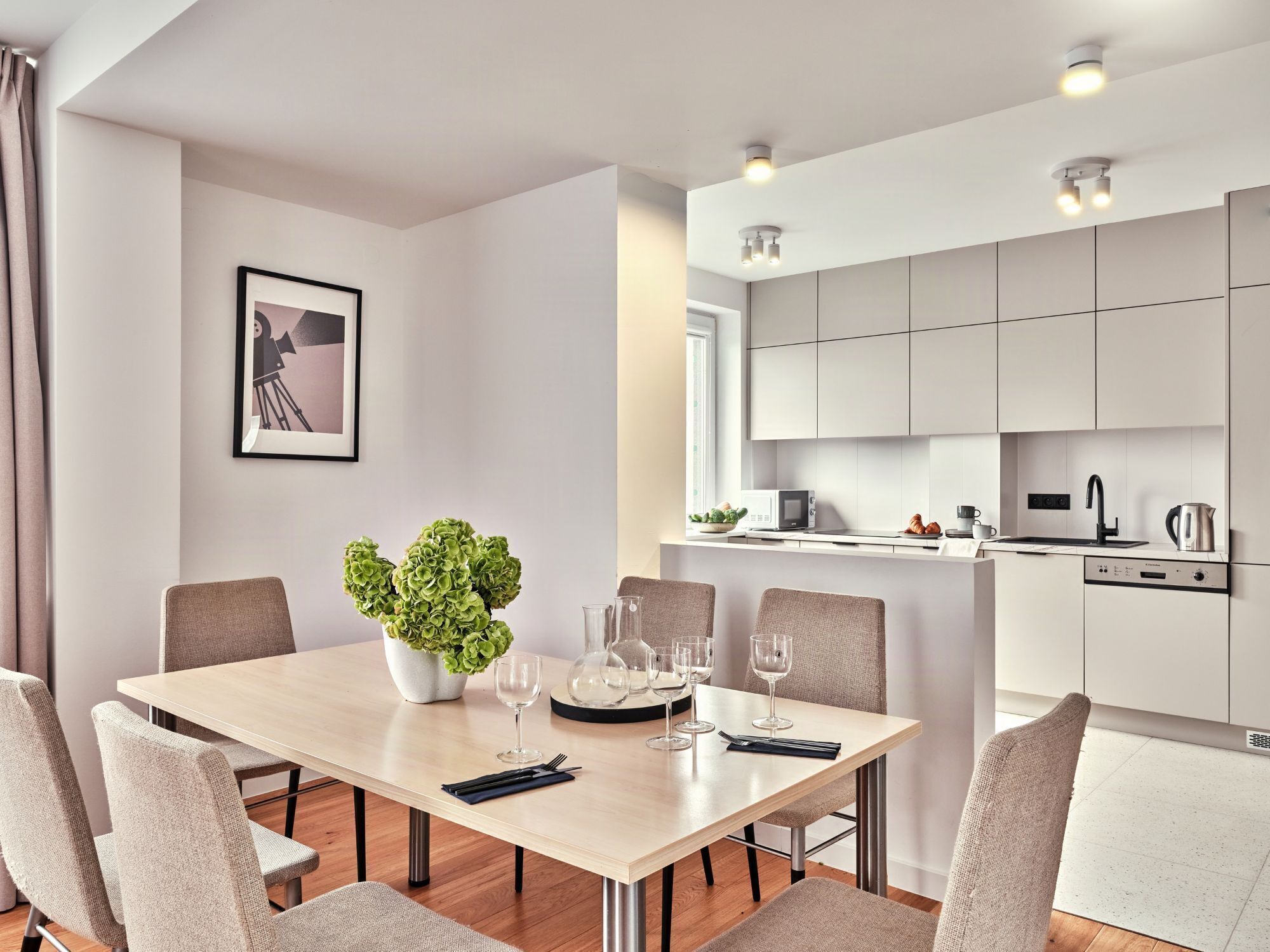 Una cocina moderna con armarios blancos, una zona de comedor con una mesa dispuesta para cuatro personas, y una planta en maceta sobre la mesa.