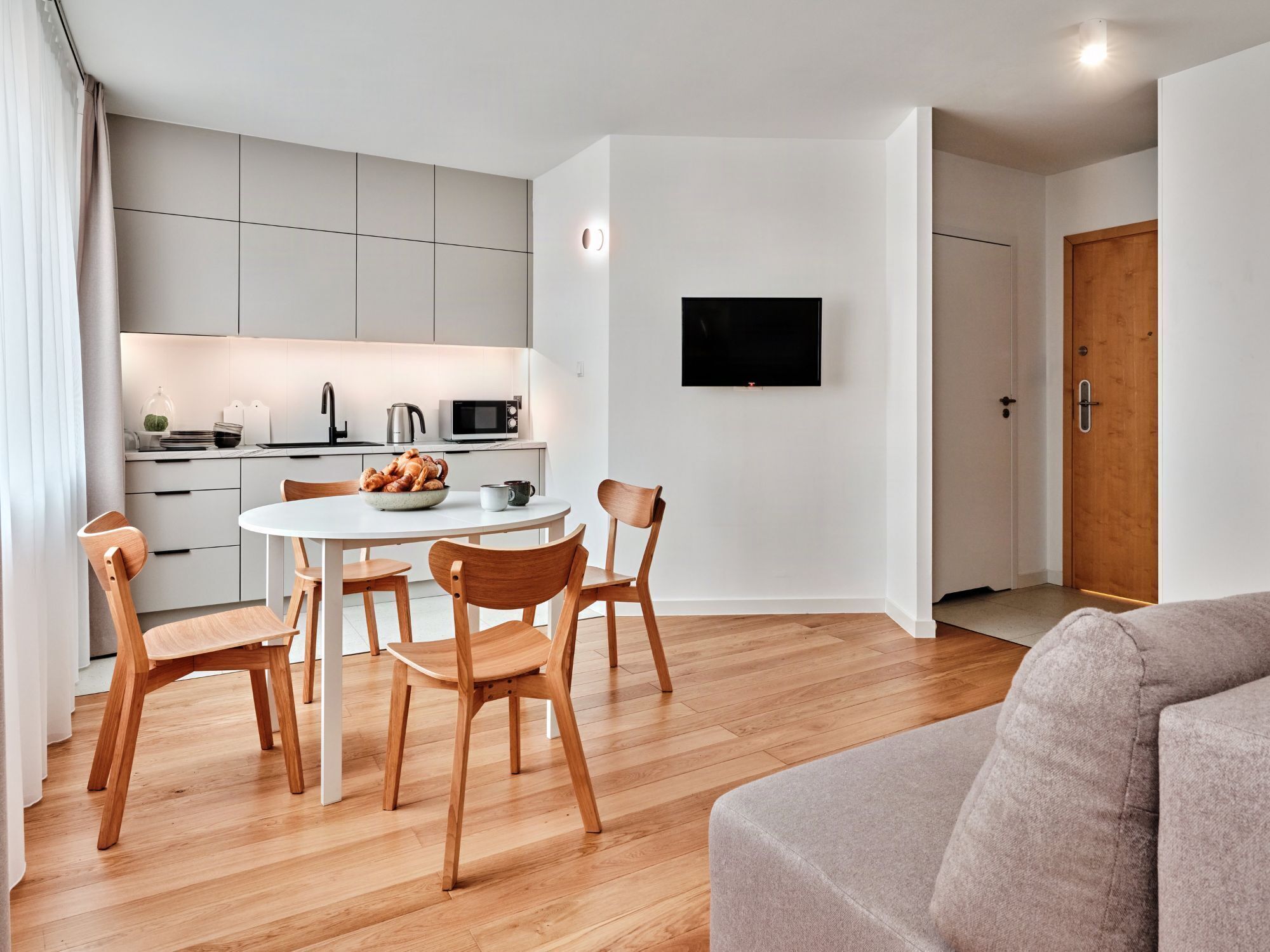 A modern kitchen with white cabinets, a dining table, and chairs. A flat-screen TV is mounted on the wall, and a wooden door is visible.