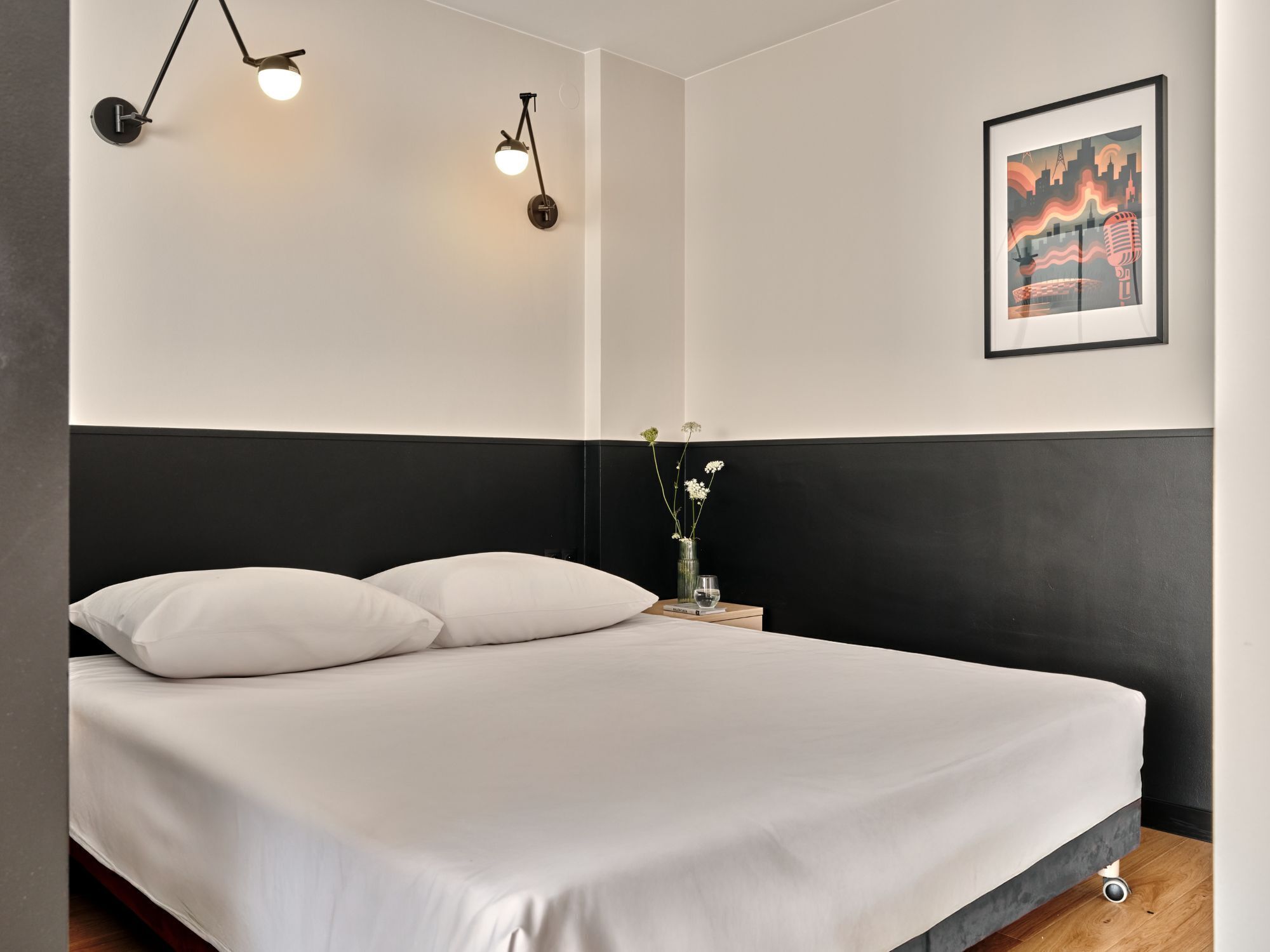 Ein modernes Schlafzimmer mit einem Bett, weißer Bettwäsche, einem schwarzen Kopfteil, einem Nachttisch mit einer Vase und einem Glas sowie einem gerahmten Bild an der Wand.