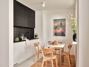A modern kitchen with white cabinets, a black countertop, and a dining area with wooden chairs and a table. - Media Park (Warsaw)