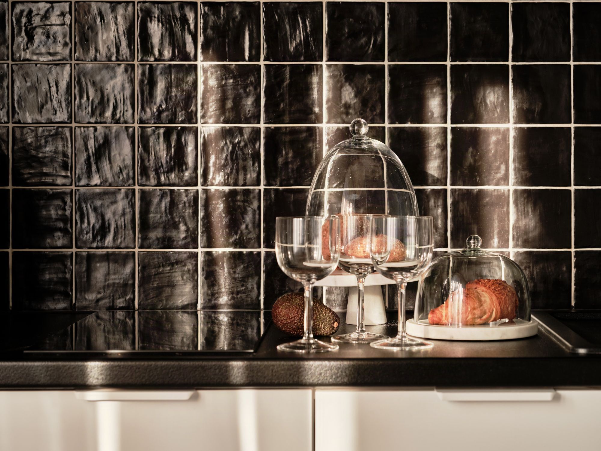 A kitchen counter with a black tiled backsplash, two wine glasses, and a glass dome covering a loaf of bread.