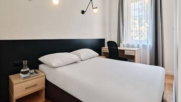 A hotel room with a bed, bedside table, lamp, desk, chair, and a window with curtains.