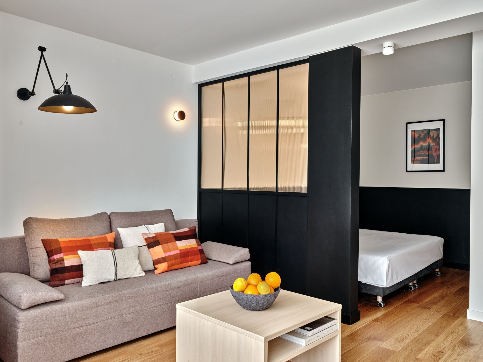 A modern living room with a sofa, a coffee table with a bowl of fruit, a bed, and a wall-mounted lamp.