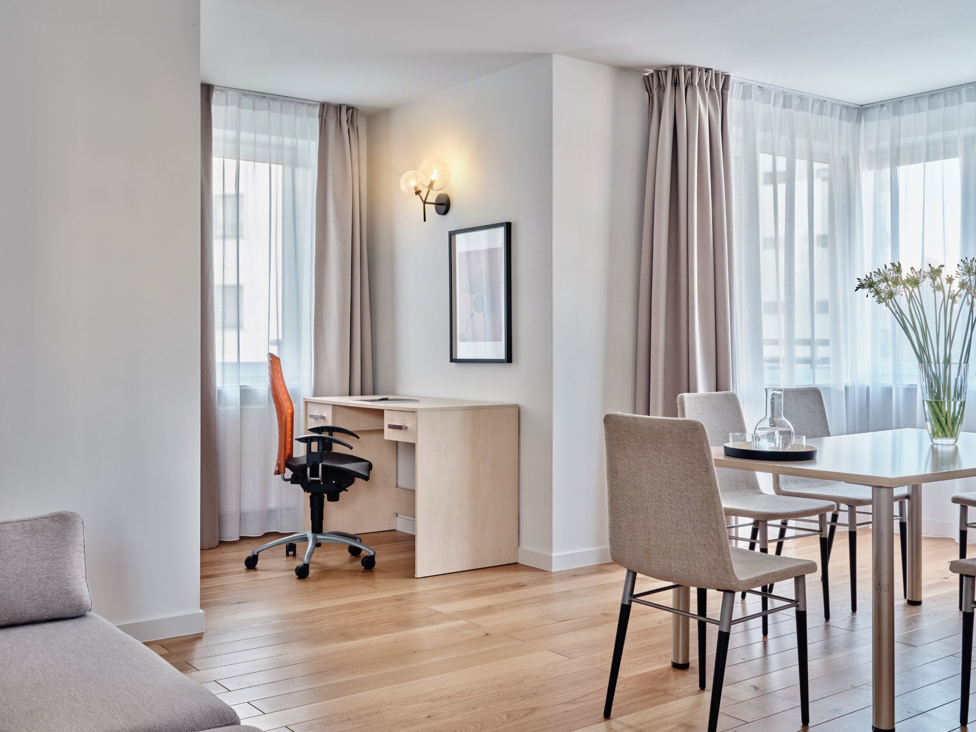 Een moderne hotelkamer met een houten vloer, een bureau met stoel, een eettafel met stoelen, en een vaas met bloemen.