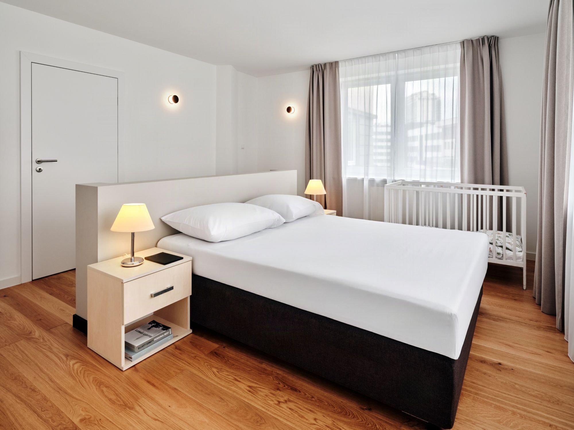 Una moderna camera d'hotel con un letto grande, lampade da comodino, un comodino con un libro e vista sulla città attraverso tende leggere.