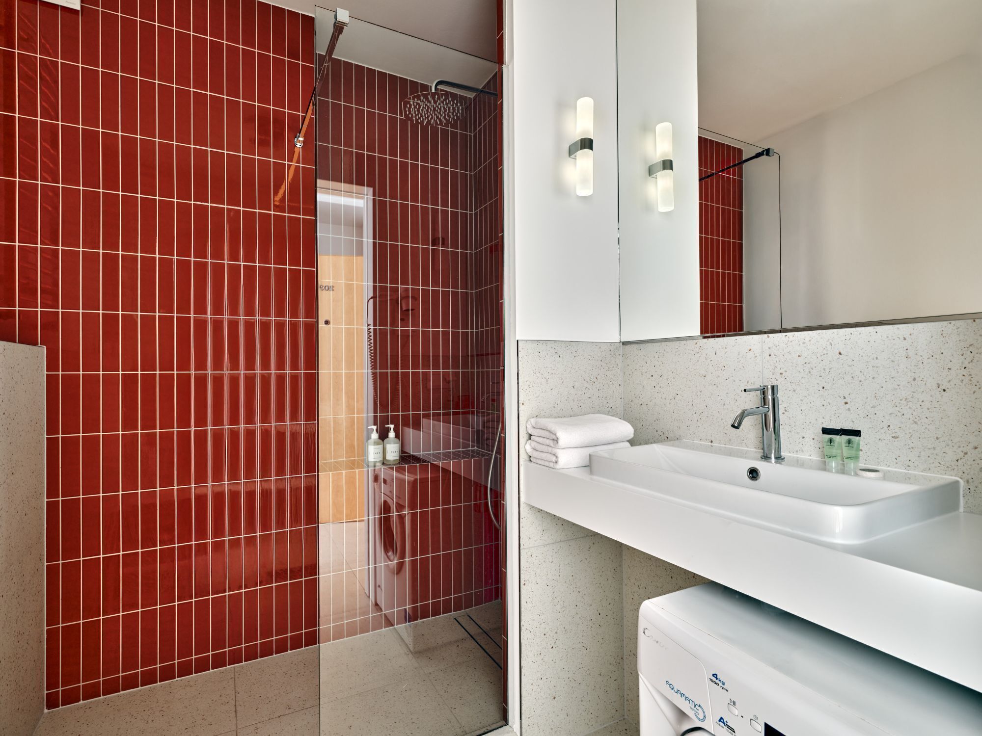 A modern bathroom with red tiled walls, a shower, a white sink, and a mirror.