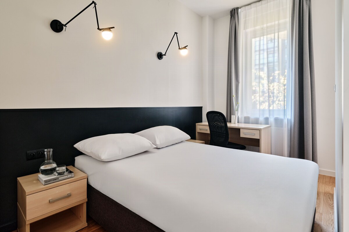 A hotel room with a bed, bedside table, lamp, desk, chair, and window with curtains.
