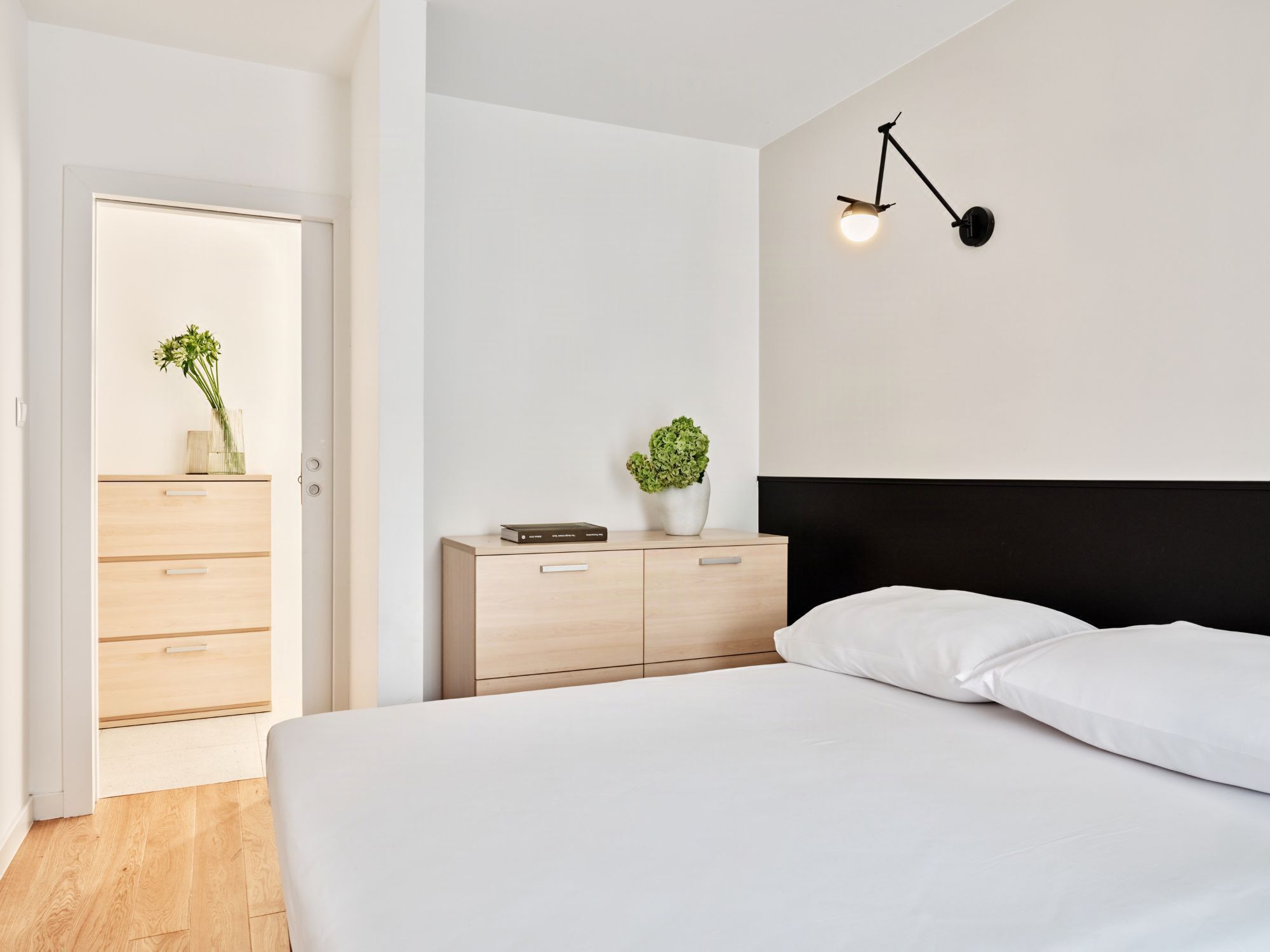 A bedroom with a bed, a bedside table with a plant, a wall-mounted lamp, and a wooden wardrobe.