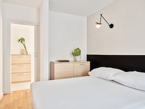 A bedroom with a bed, a bedside table with a plant, a wall-mounted lamp, and a wooden wardrobe. - Media Park (Warsaw)