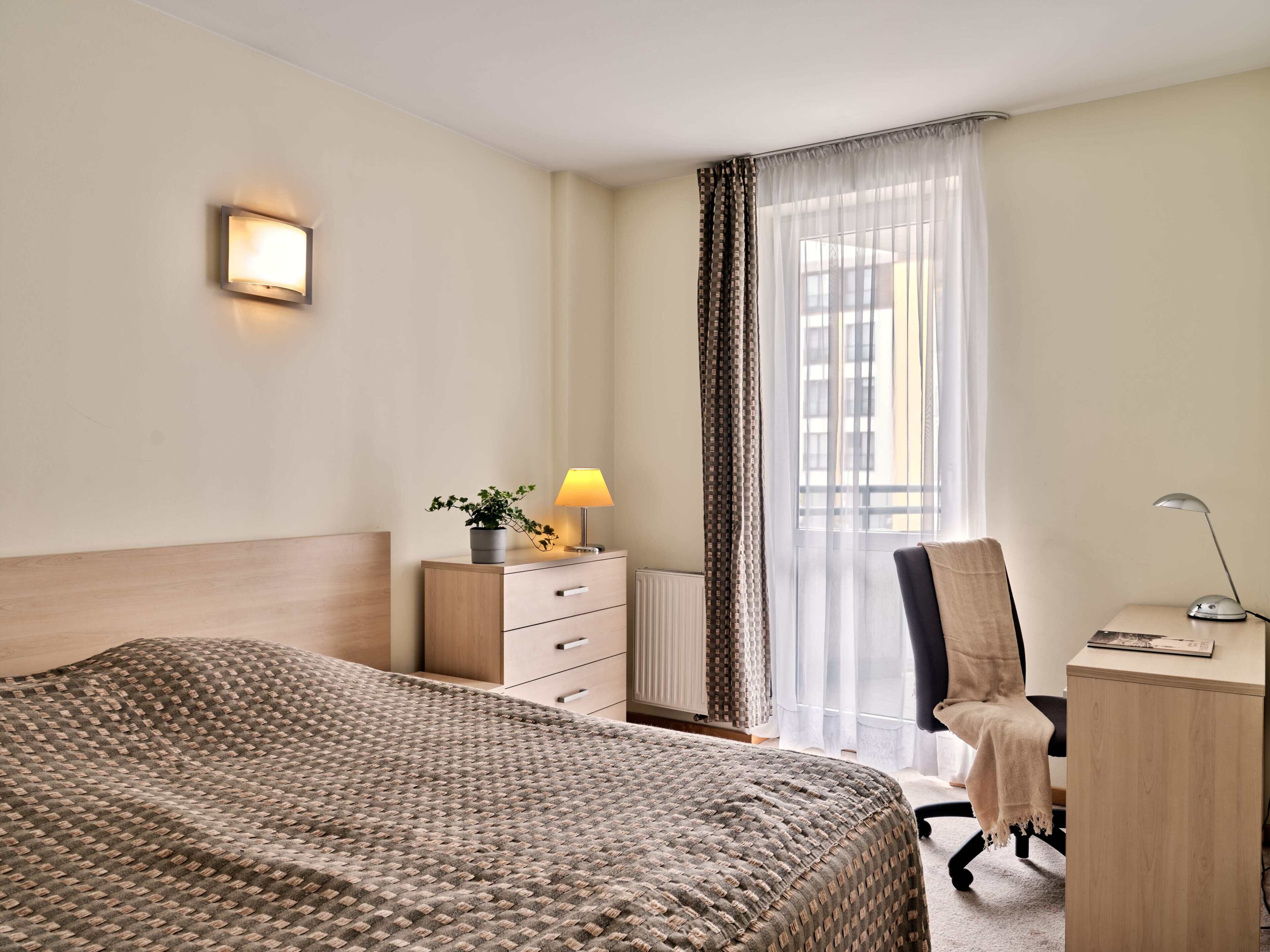 Habitación de hotel con una cama grande, un escritorio, una silla, una lámpara, una planta en maceta y una ventana con cortinas.