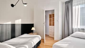 A hotel room with two beds, a bedside table with a lamp, a wardrobe, and a door leading to a bathroom.