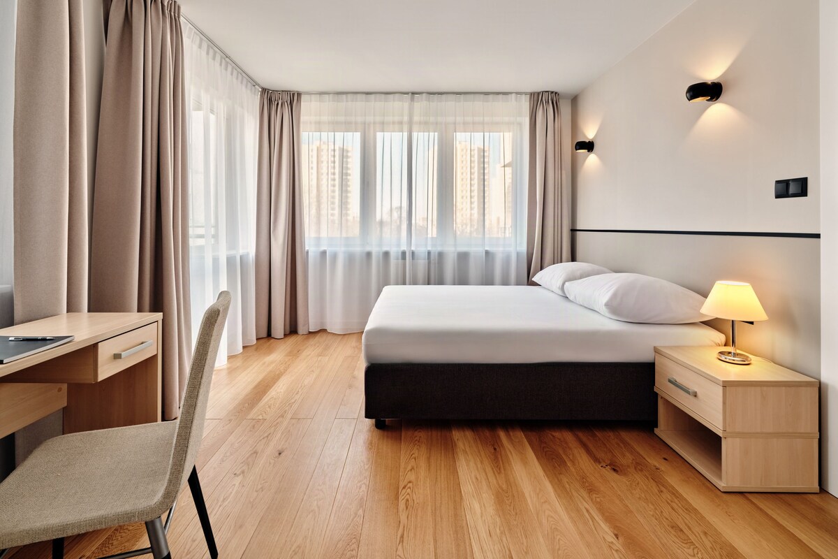 A hotel room with a bed, bedside table, lamp, chair, desk, and curtains.