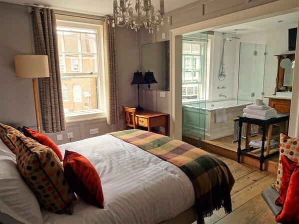 A hotel room with a large bed, a wooden floor, a desk, a chair, a lamp, a mirror, and a bathroom with a glass shower.