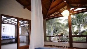 A room with a bed, a dining area with chairs, a table with flowers, and large windows with a view of palm trees. - Encantos da Ilha Pousada (Florianópolis)
