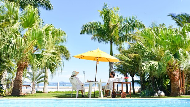 A poolside area with a table and chairs under a yellow umbrella, surrounded by palm trees and a view of the sea. - Encantos da Ilha Pousada (Florianópolis)
