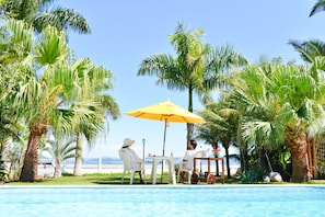 A poolside area with a table and chairs under a yellow umbrella, surrounded by palm trees and a view of the sea.