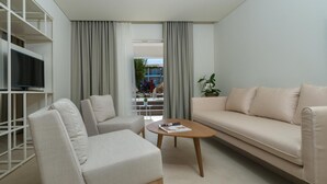 Un salon moderne avec un canapé, un fauteuil et une table basse. On y trouve un téléviseur posé sur un meuble blanc, et une plante sur la droite.