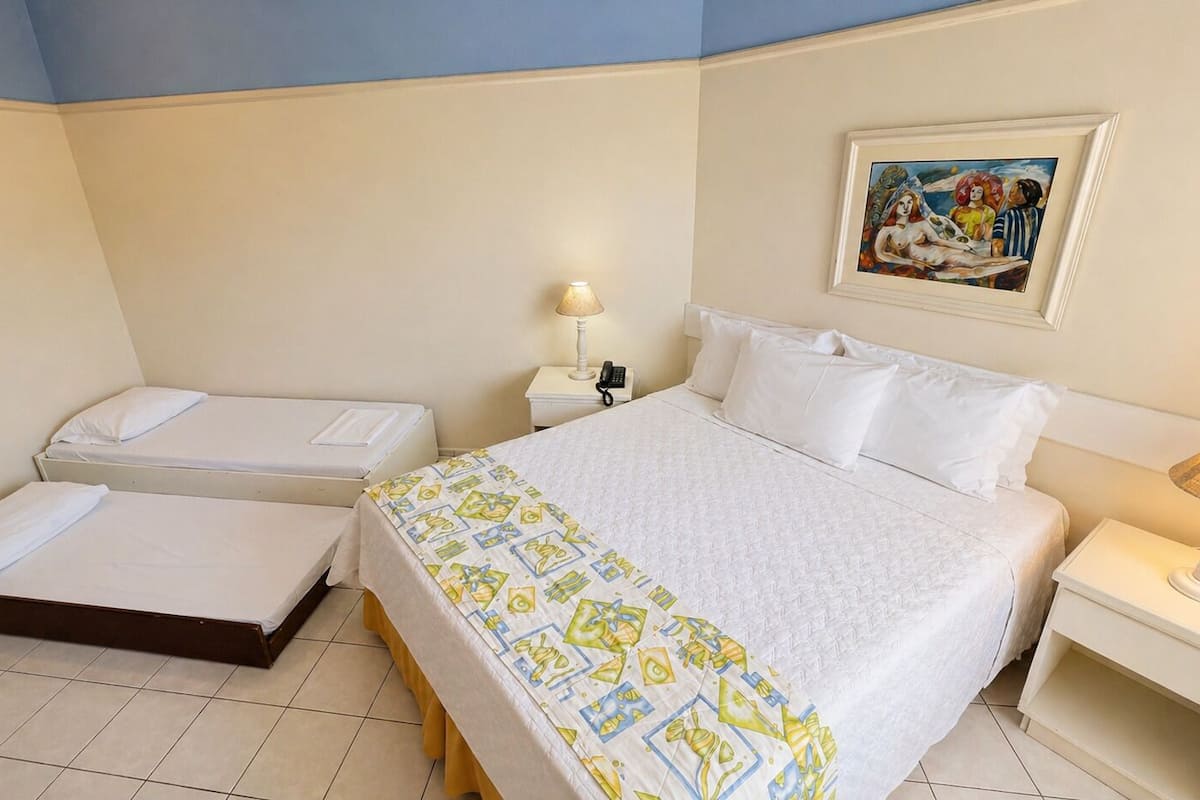 Une chambre d’hôtel avec un grand lit, deux petits lits, une table de chevet avec une lampe, une commode et un tableau au mur.