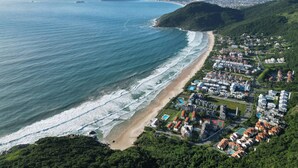 An aerial view of a coastal resort with a beachfront, dense residential area, and hilly terrain. - Hotel Praia Brava (Florianópolis)