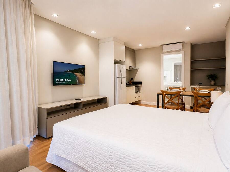 A hotel room with a bed, a desk, a dining table, and a TV displaying a beach scene.