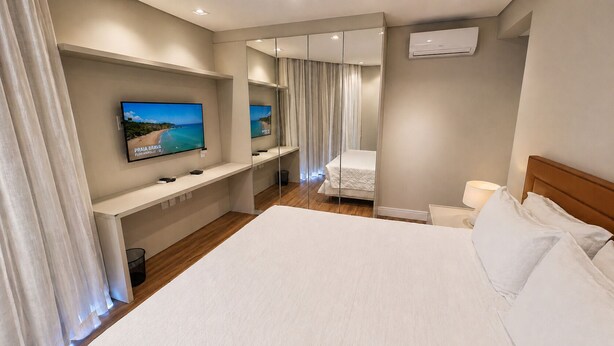 A hotel room with a large bed, a TV mounted on a wall, a mirror, and a window with curtains. - Hotel Praia Brava (Florianópolis)