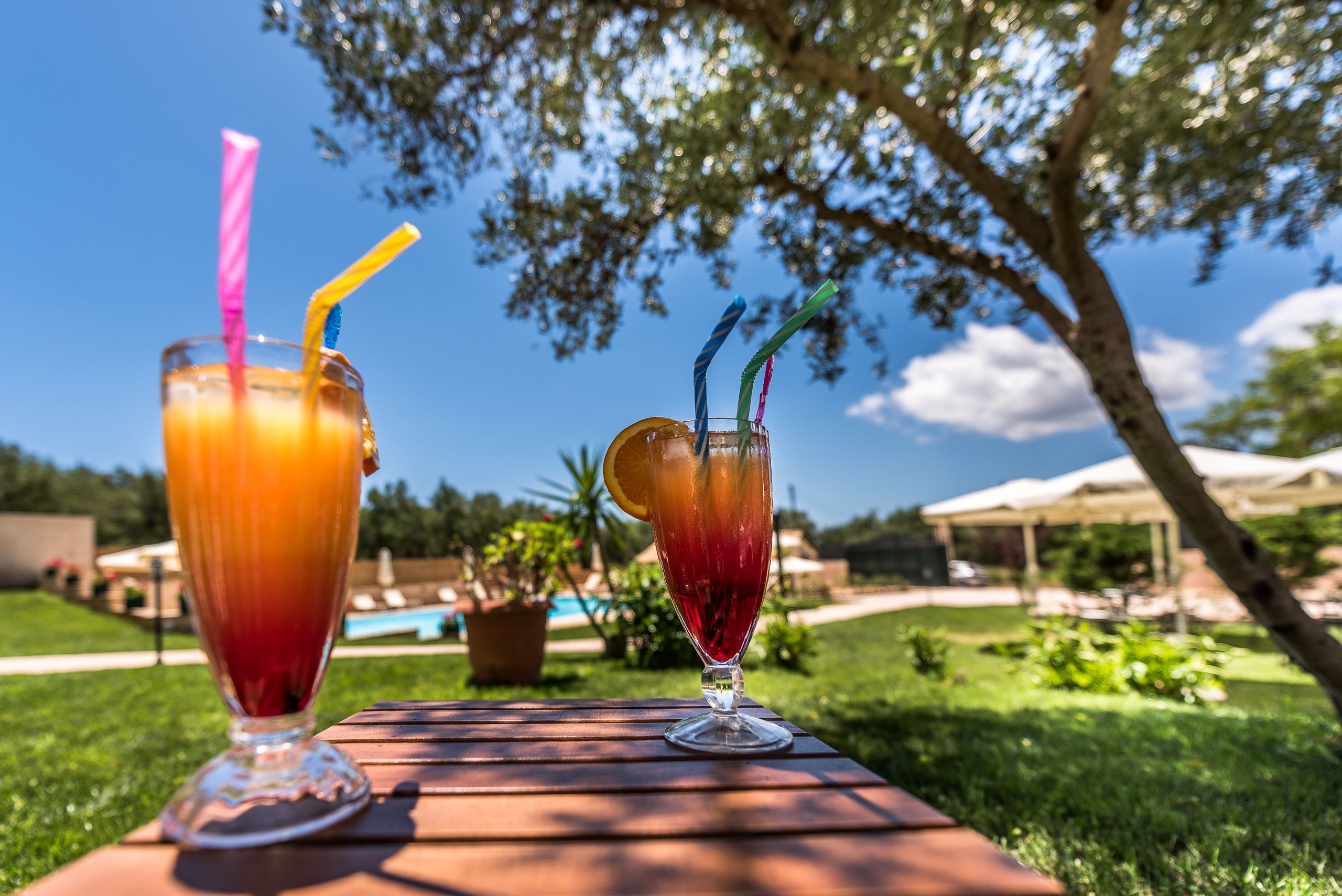 Two drinks on a wooden table outdoors with a pool and umbrellas in the background.