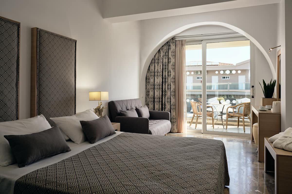 A modern hotel room with a large bed, a seating area with a table and chairs, and a balcony with a view of the building.