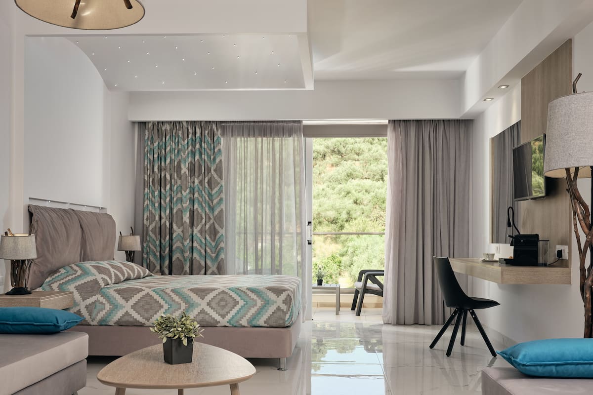 A modern hotel room with a bed, a sofa, a small table with a plant, a desk with a chair, and a balcony with a view of greenery.