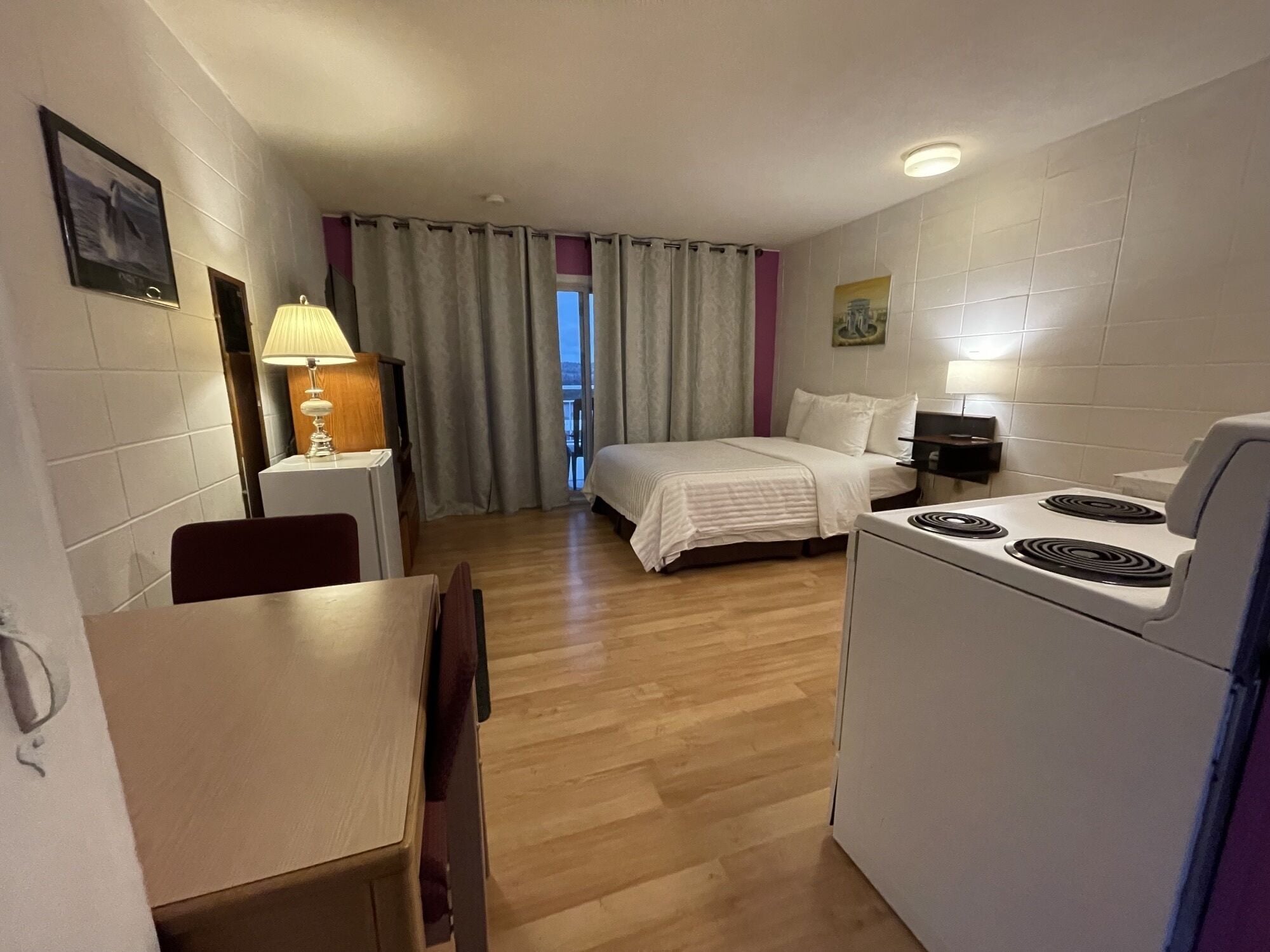 Habitación de hotel pequeña con cama, una mesita de comedor, una silla, una lámpara, cocina y vistas al exterior a través de la ventana.