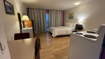 Habitación de hotel pequeña con cama, una mesita de comedor, una silla, una lámpara, cocina y vistas al exterior a través de la ventana.