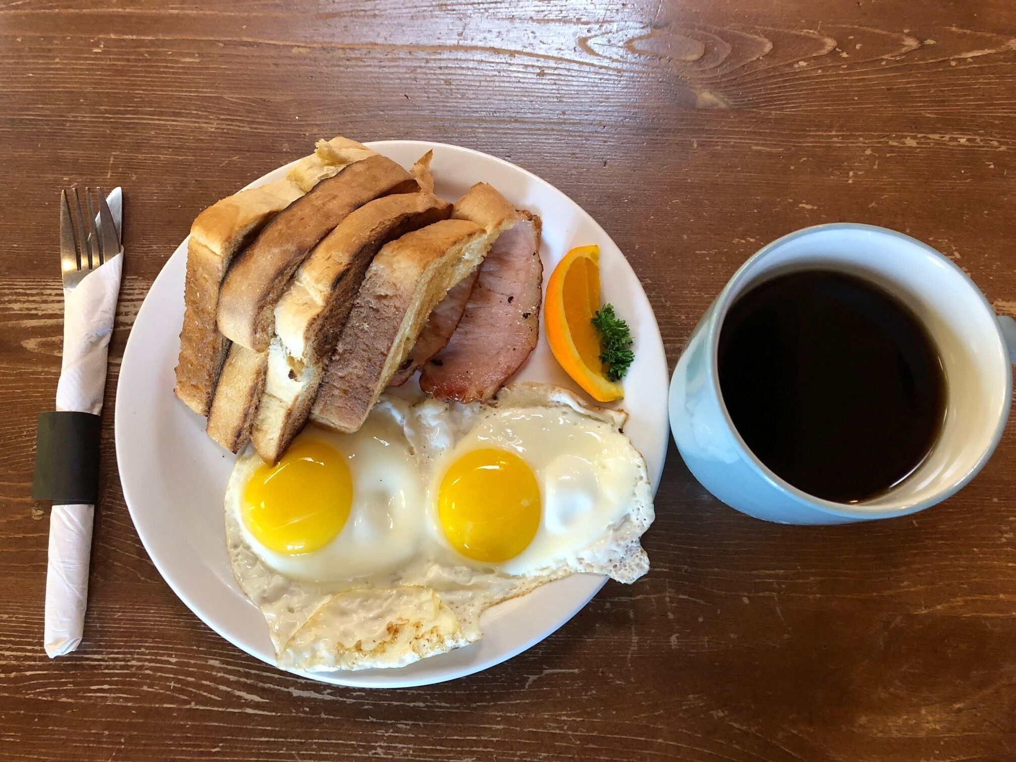 A plate with sliced meat, two fried eggs, a slice of orange, and a cup of coffee on a wooden table.